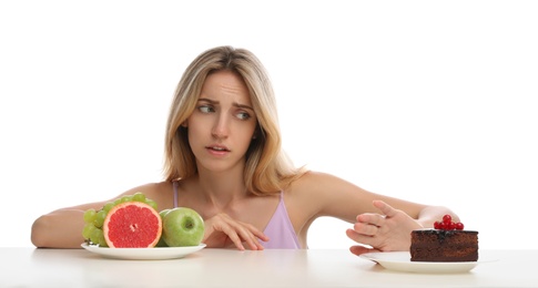 Woman choosing between cake and healthy fruits at table on white background