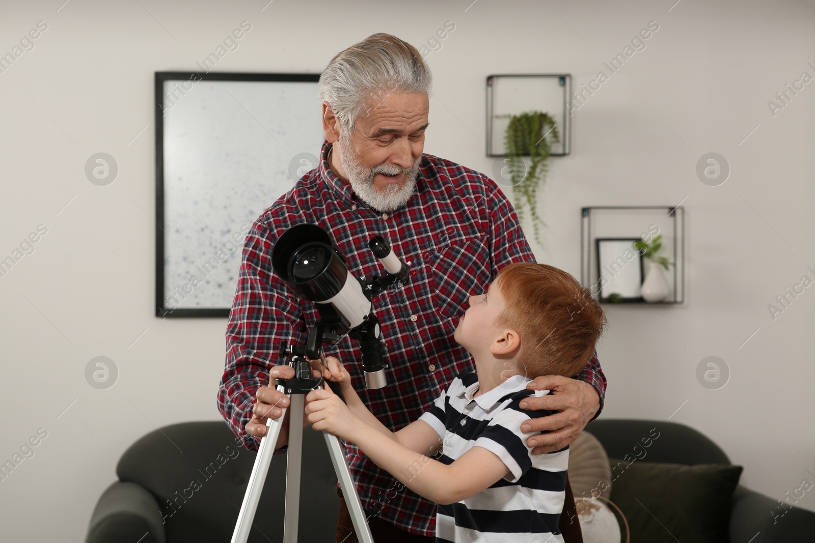 Photo of Little boy with his grandfather using telescope in room at home