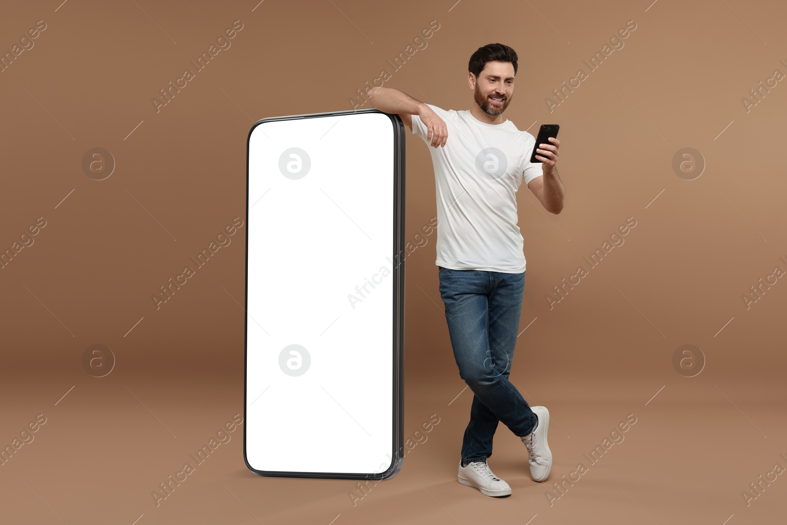 Image of Man with mobile phone standing near huge device with empty screen on dark beige background. Mockup for design