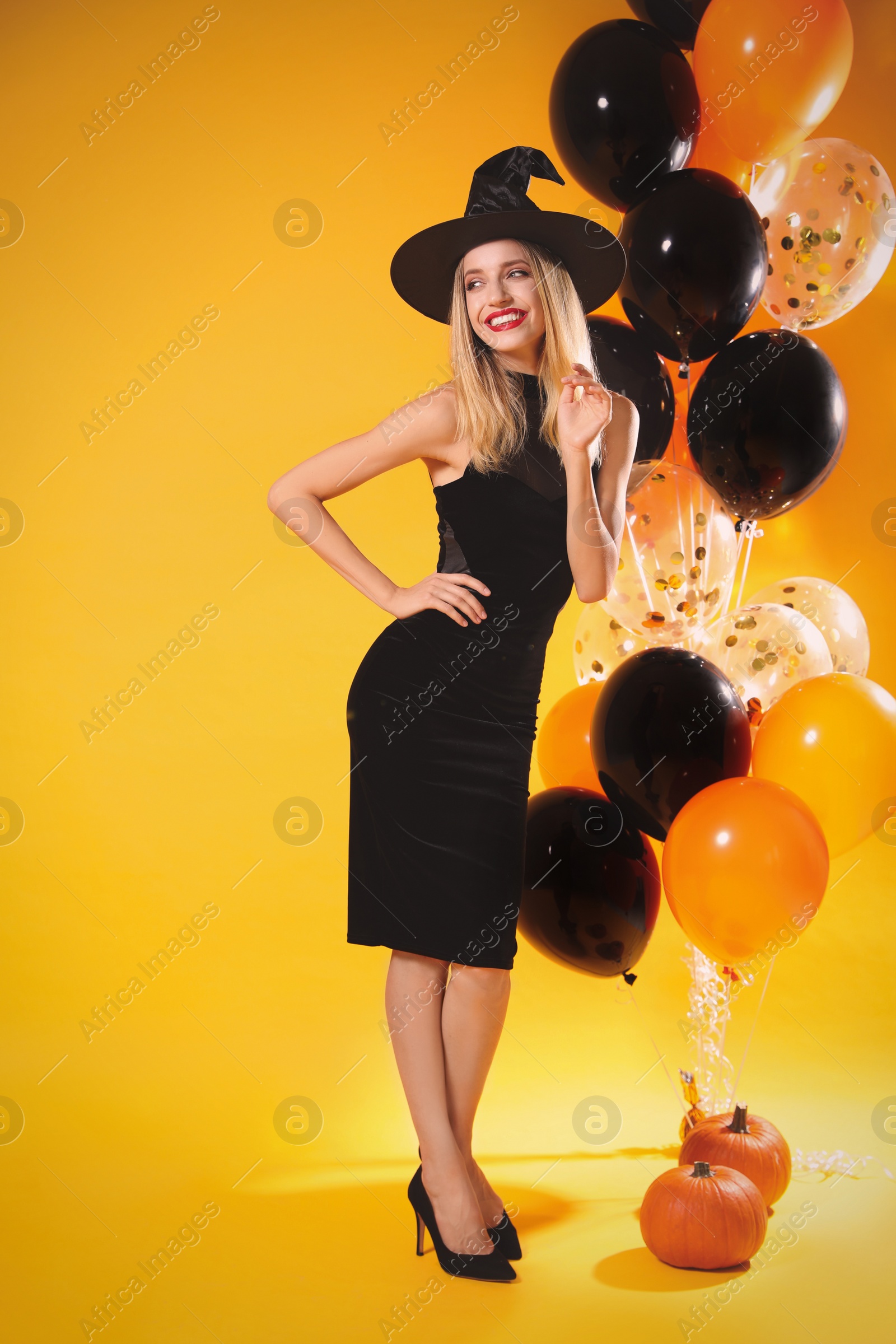 Photo of Beautiful woman in witch costume with balloons and pumpkins on yellow background. Halloween party