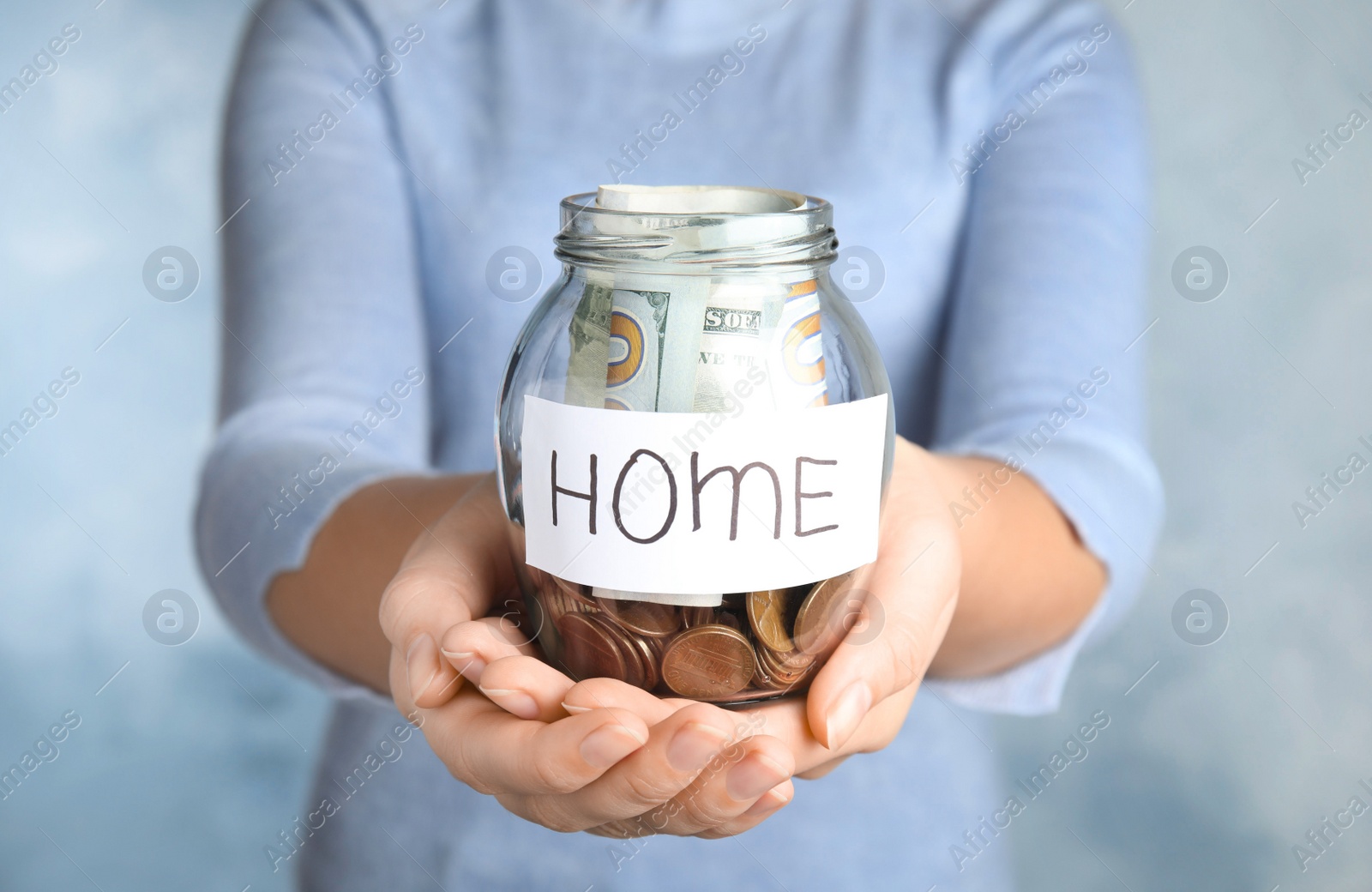 Photo of Woman holding glass jar with money and tag HOME on light blue background, closeup