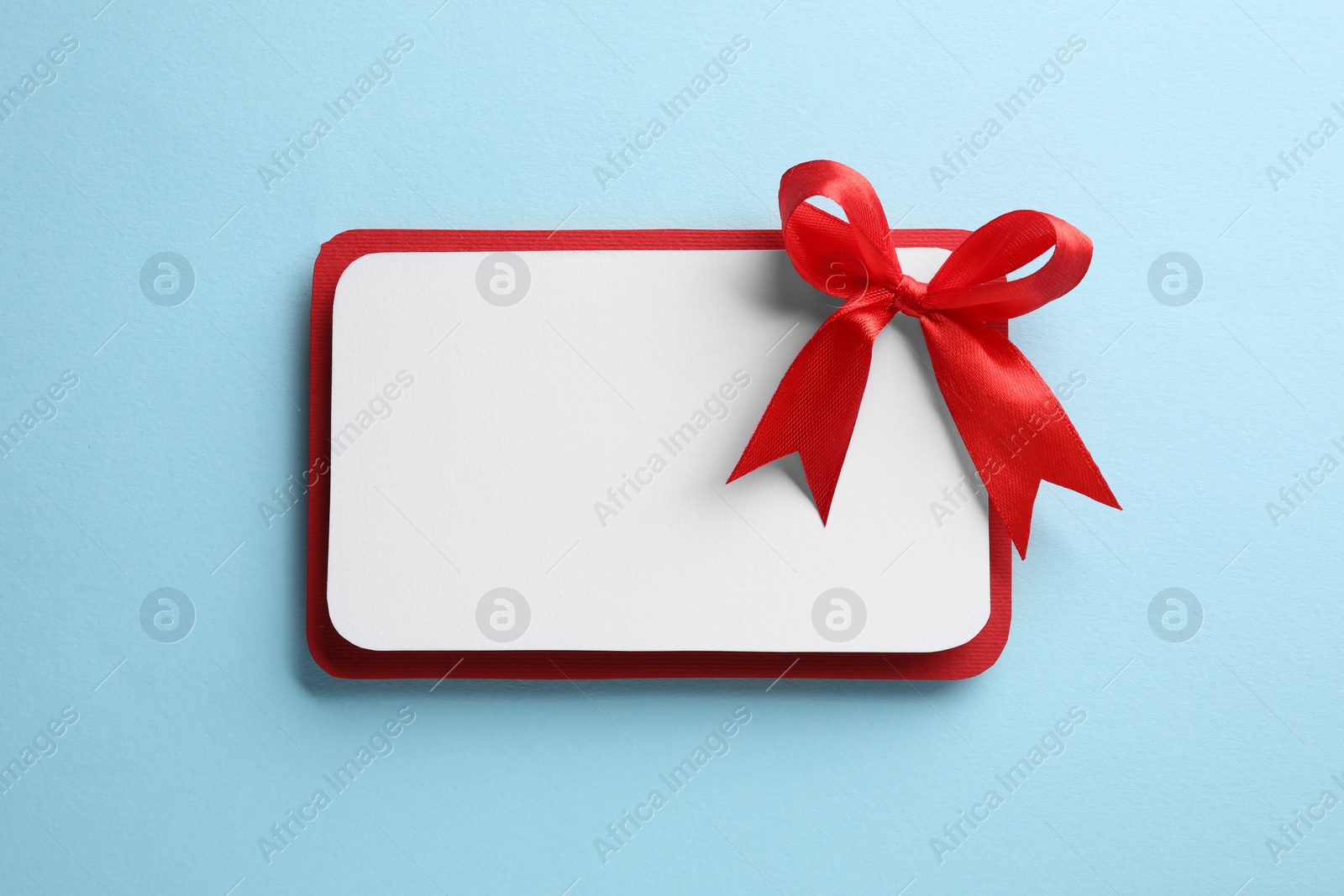 Photo of Blank gift card with red bow on light blue background, top view