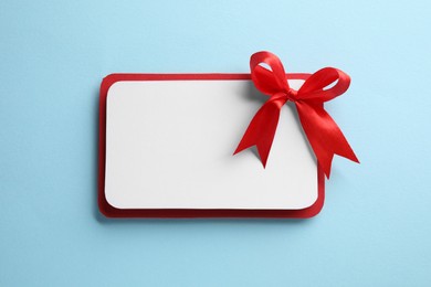 Photo of Blank gift card with red bow on light blue background, top view