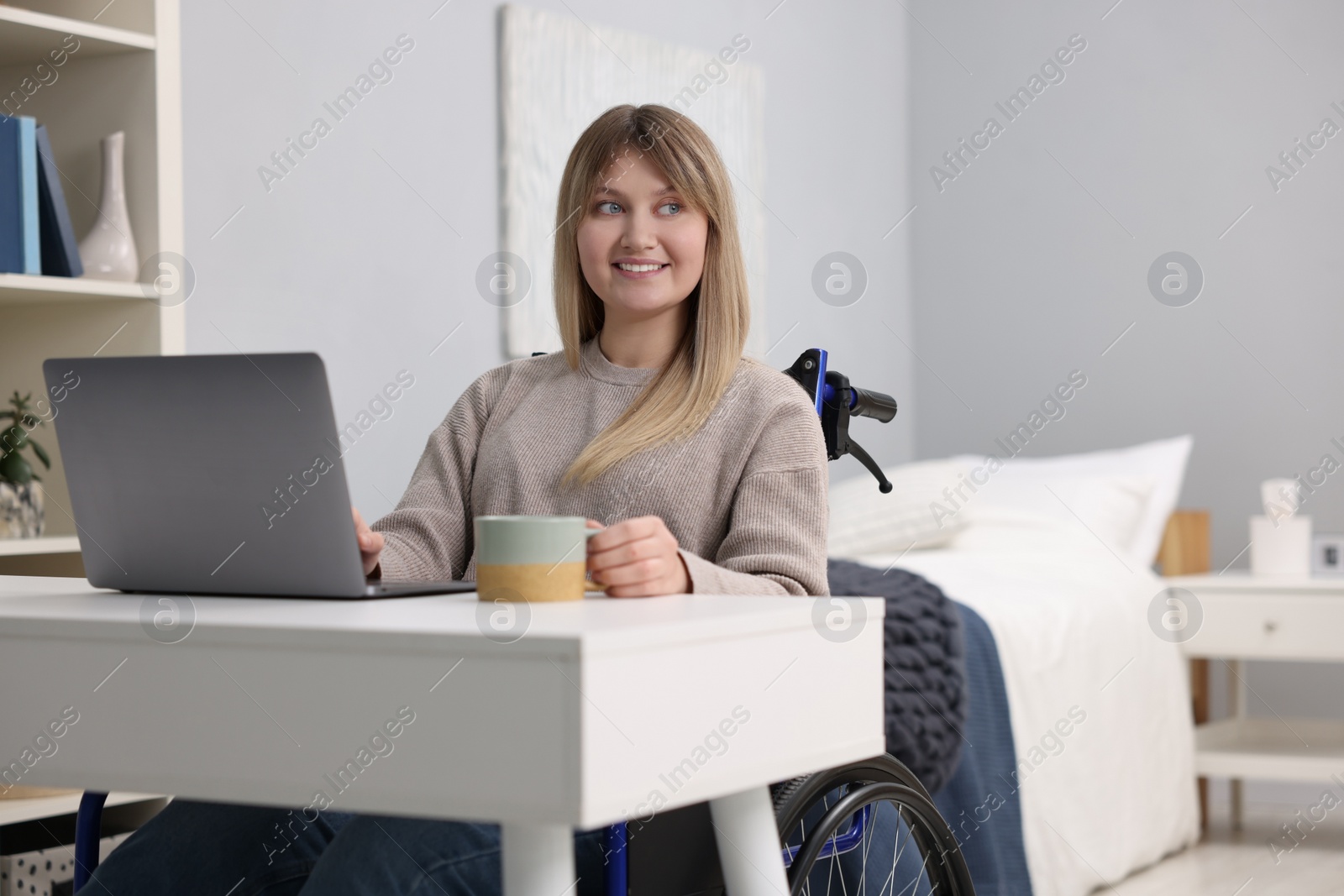 Photo of Woman in wheelchair with cup of drink using laptop at table in home office