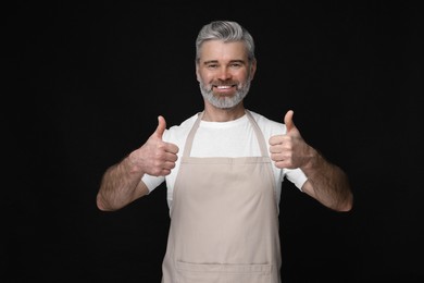 Happy man in kitchen apron showing thumbs up on black background. Mockup for design