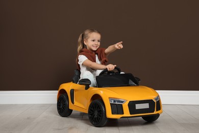 Cute little girl driving children's electric toy car near brown wall indoors