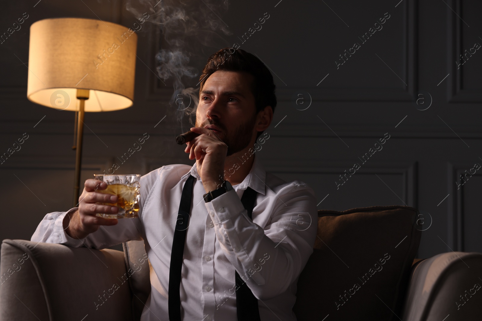 Photo of Handsome man with glass of whiskey smoking cigar at home. Space for text