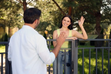 Photo of Friendly relationship with neighbours. Happy woman greeting man near fence outdoors