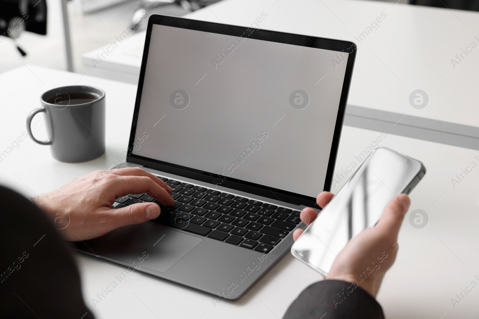 Photo of Man using phone while working on laptop at white desk in office, closeup