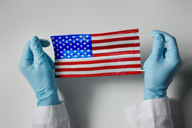 Image of Doctor holding medical mask with USA flag pattern on light grey background, closeup. Dangerous virus