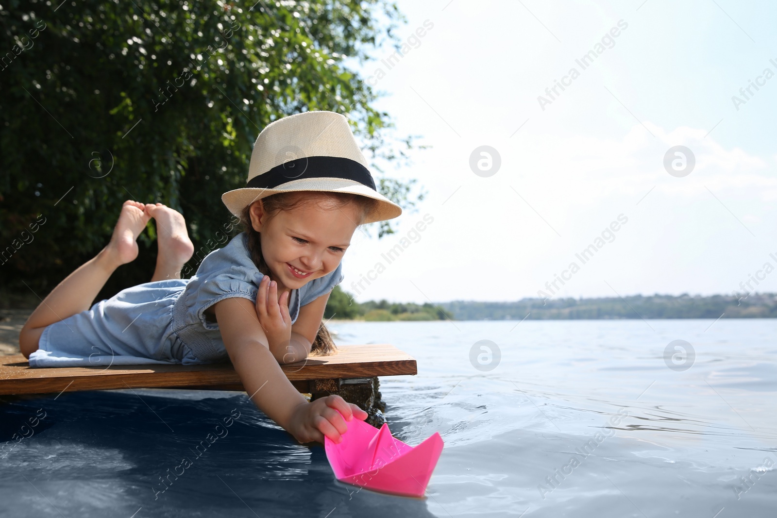 Photo of Cute little girl playing with paper boat on wooden pier near river