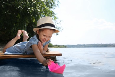 Cute little girl playing with paper boat on wooden pier near river