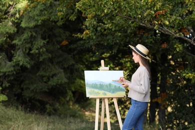 Young woman drawing on easel in forest, space for text