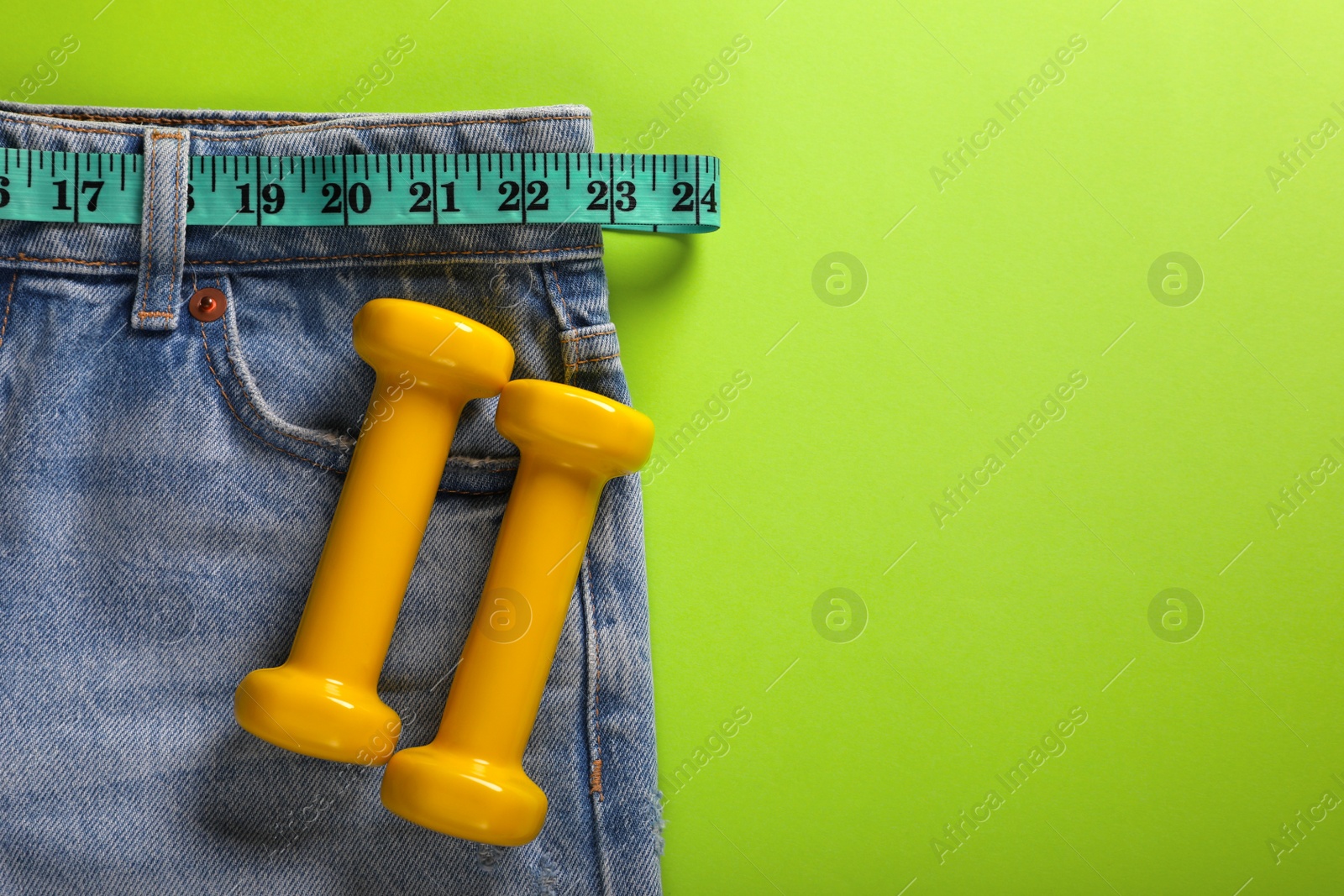 Photo of Jeans, dumbbells and measuring tape on light green background, flat lay with space for text. Weight loss concept