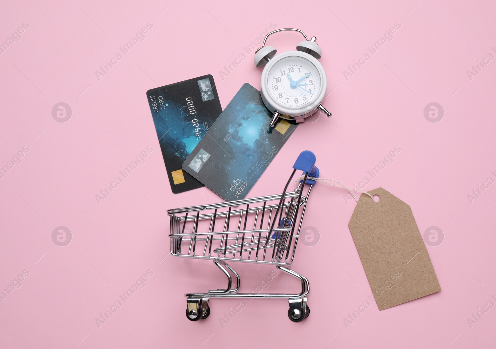 Photo of Shopping cart with tag, credit cards and alarm clock on pink background, flat lay