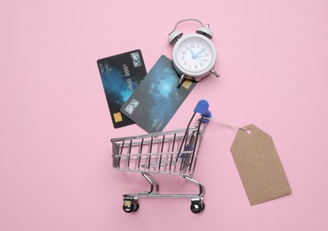 Shopping cart with tag, credit cards and alarm clock on pink background, flat lay