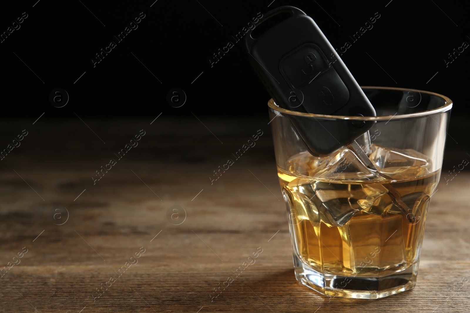 Photo of Car key in glass of alcohol on wooden table, space for text. Dangerous drinking and driving