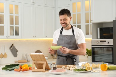 Photo of Happy man making dinner while watching online cooking course via tablet in kitchen