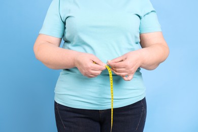 Photo of Overweight woman measuring waist with tape on light blue background, closeup