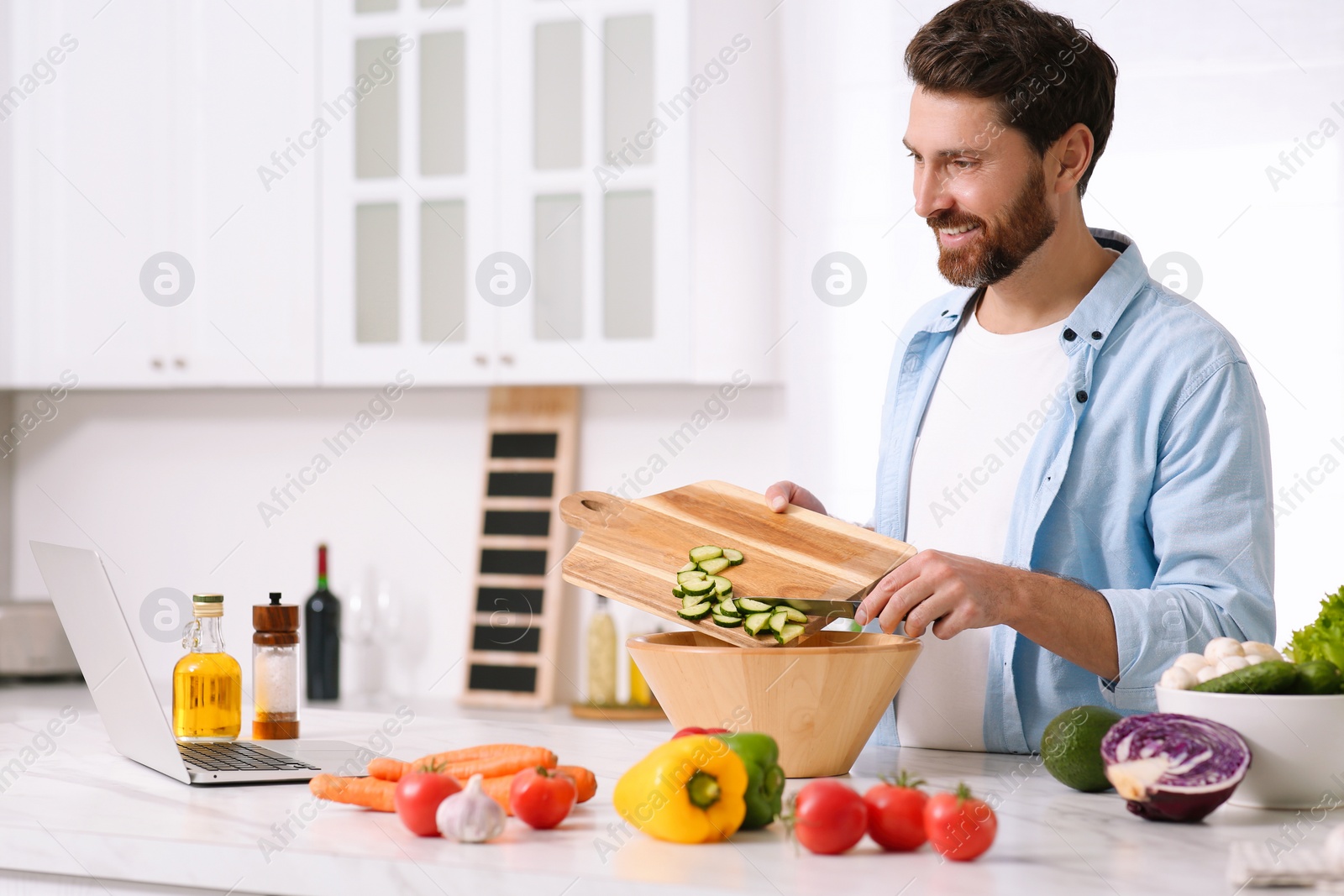 Photo of Man making dinner while watching online cooking course via laptop in kitchen