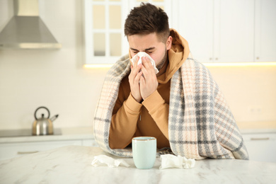 Photo of Sick young man with cup of hot drink and tissues in kitchen. Influenza virus