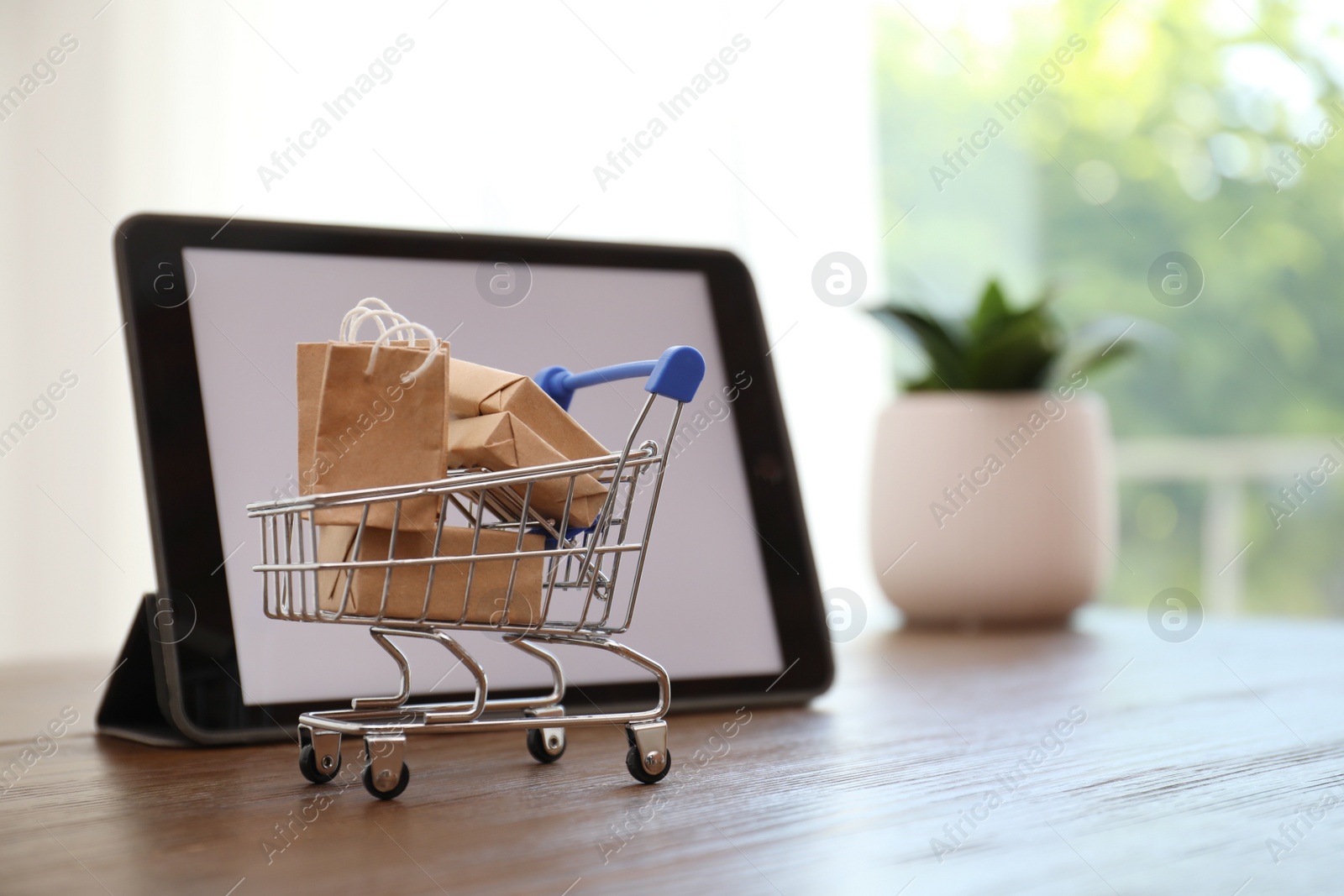 Photo of Internet shopping. Small cart with bags and boxes near modern tablet on table indoors, space for text