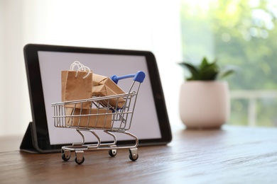 Internet shopping. Small cart with bags and boxes near modern tablet on table indoors, space for text