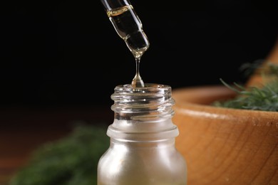 Dripping dill essential oil from pipette into bottle against black background, closeup