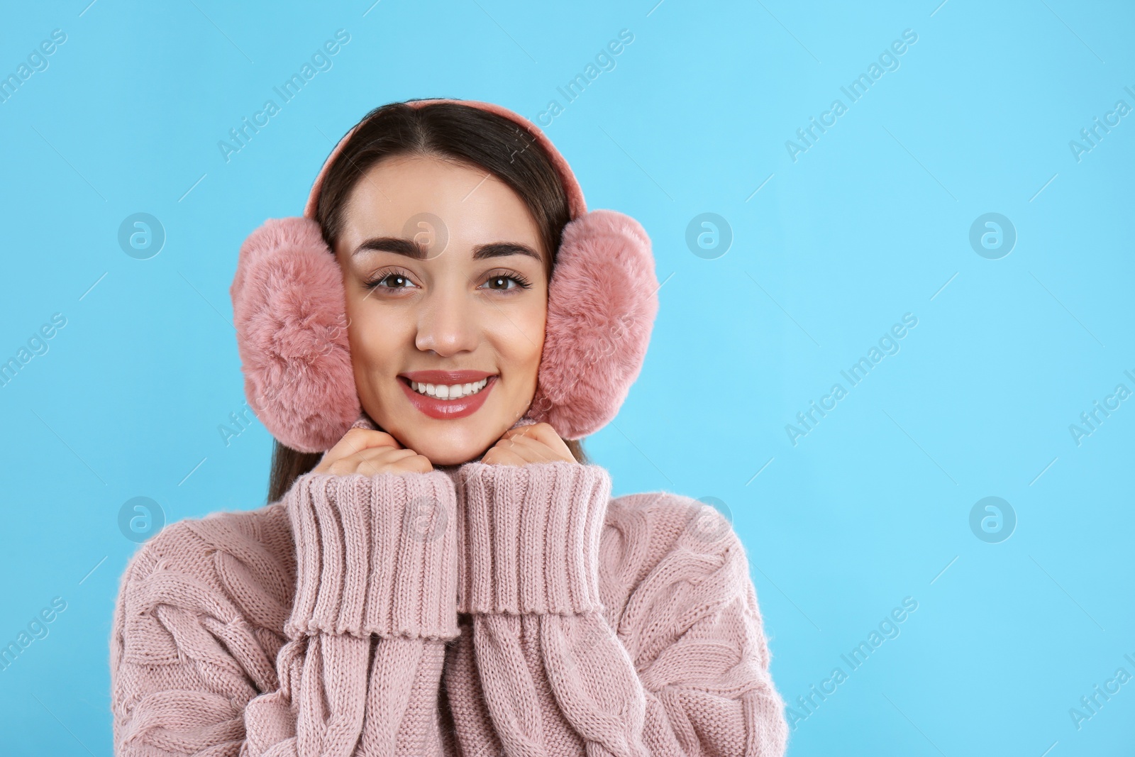 Photo of Beautiful young woman wearing earmuffs on light blue background. Space for text