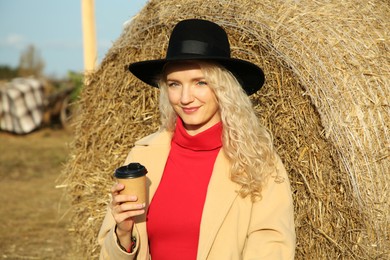 Beautiful woman with cup of hot drink sitting near hay bale outdoors. Autumn season