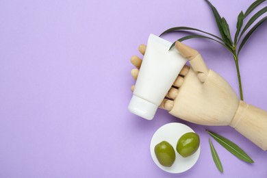 Photo of Wooden mannequin hand with tube of natural cream, olives and green leaves on violet background, flat lay. Space for text