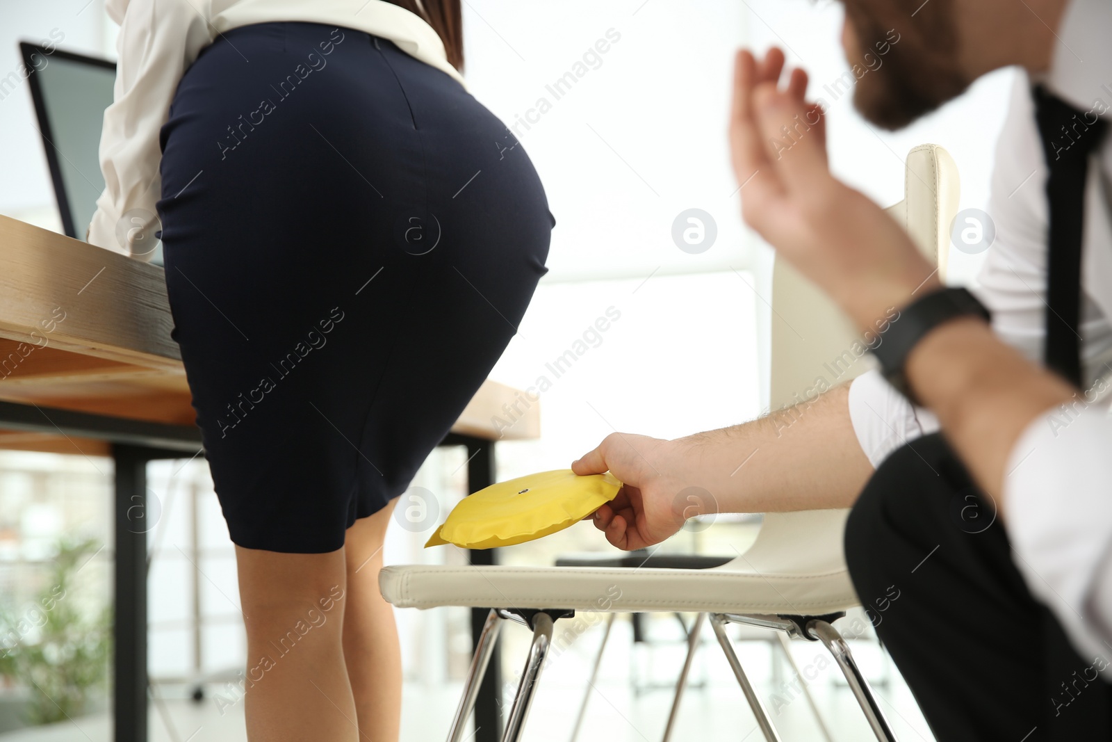 Photo of Young man putting whoopee cushion on chair while his colleague sitting down in office, closeup. Funny joke