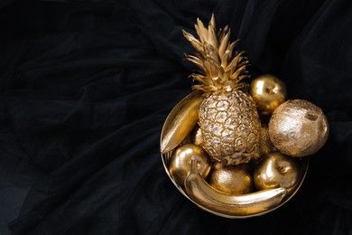 Photo of Golden bowl with fruits on black fabric, above view. Space for text