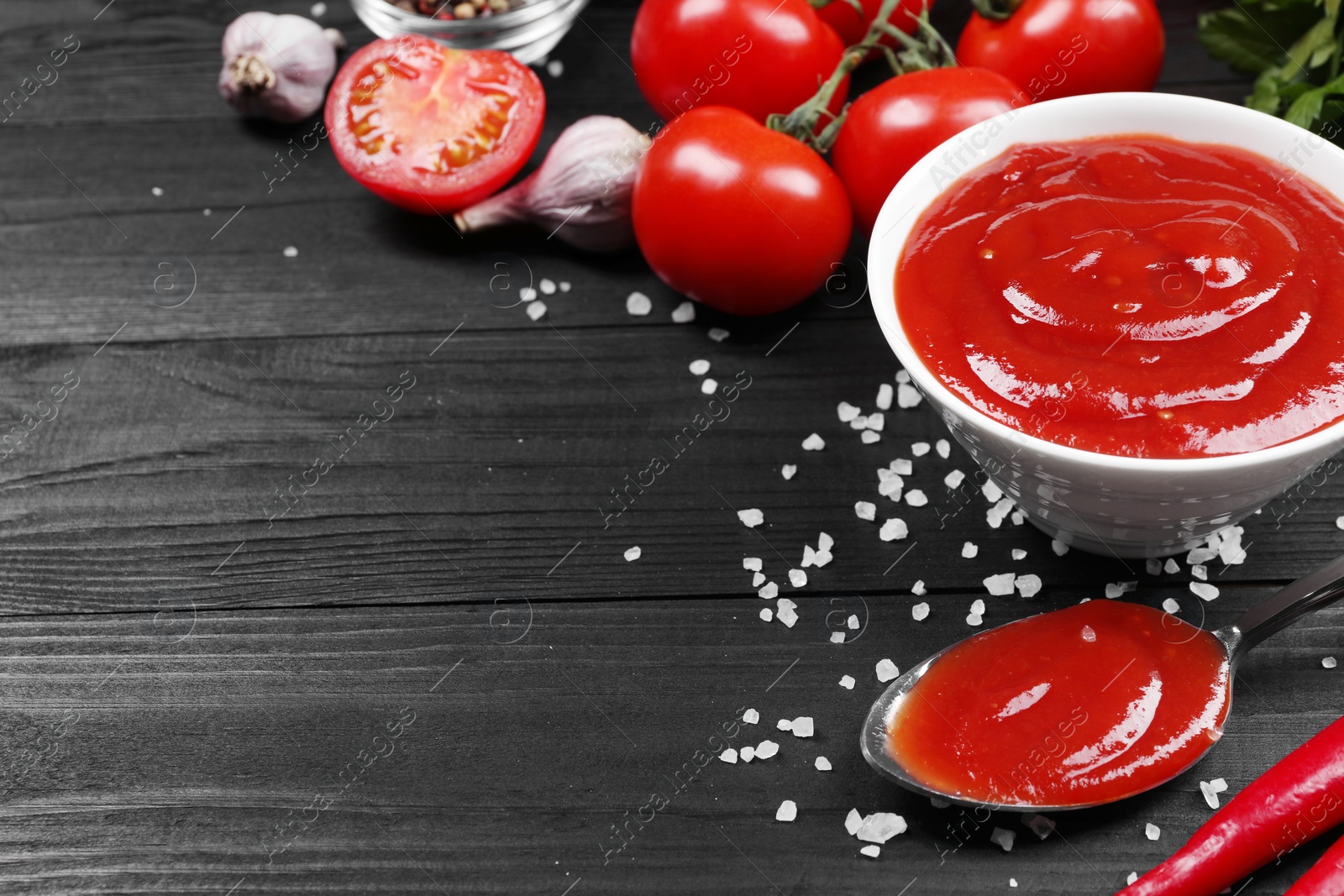 Photo of Delicious ketchup in bowl, salt and products on black wooden table, space for text. Tomato sauce