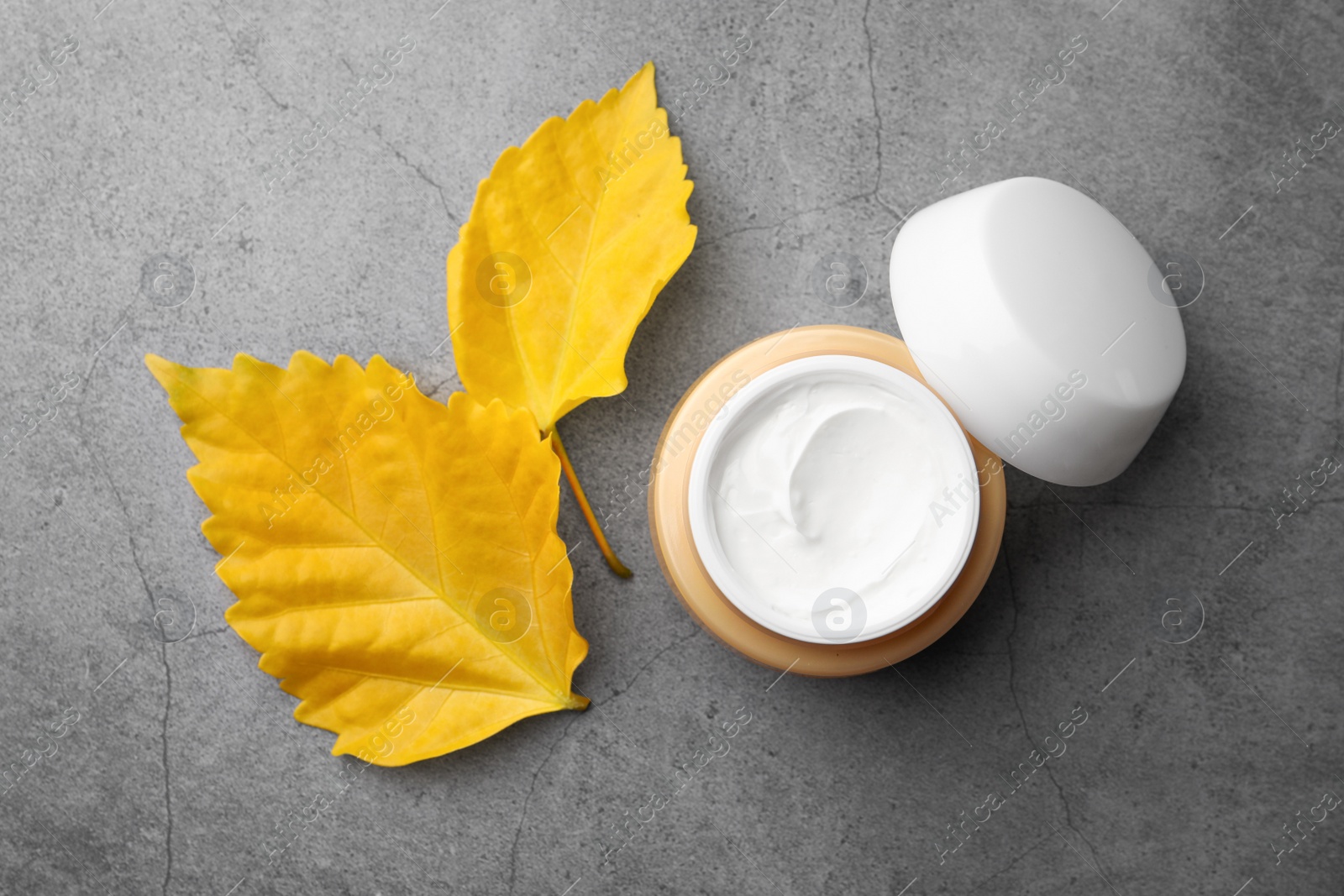 Photo of Jar of face cream and yellow leaves on grey table, top view