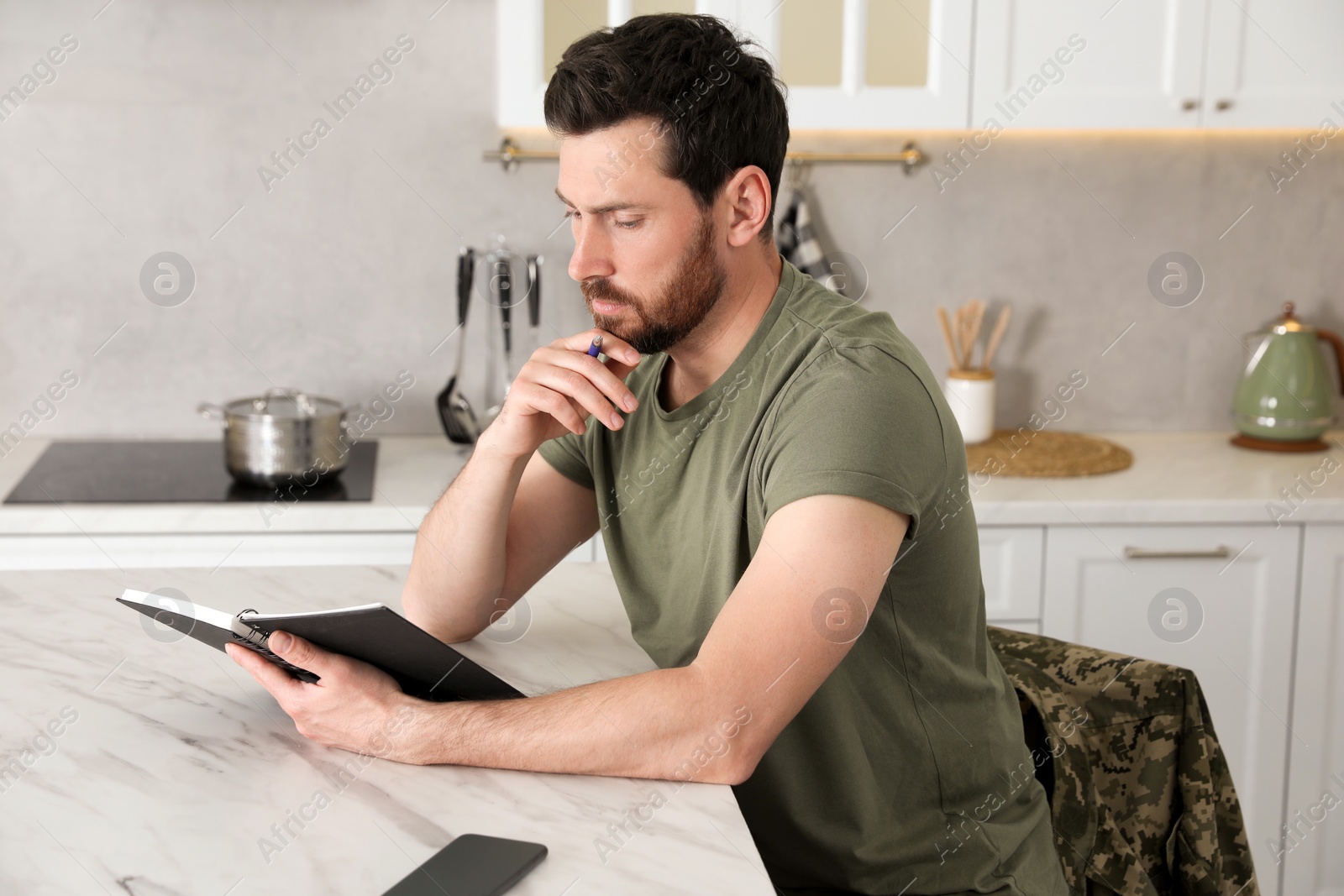 Photo of Soldier with notebook at white marble table in kitchen. Military service