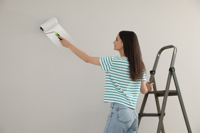 Young woman painting wall with roller on ladder indoors