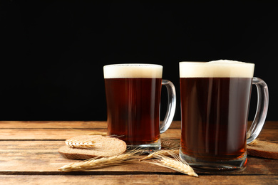 Mugs of delicious kvass, spikes and bread on wooden table against black background. Space for text