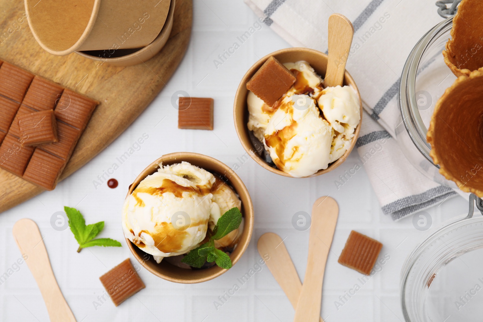 Photo of Scoops of ice cream with caramel sauce, mint leaves and candies on white tiled table, flat lay