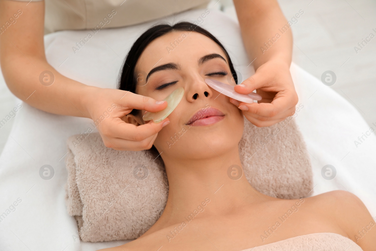 Photo of Young woman receiving facial massage with gua sha tools in beauty salon