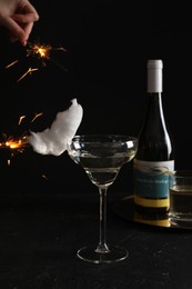 Photo of Cocktail with tasty cotton candy and bottle of alcohol drink on dark textured table. Woman holding sparkler against black background, closeup
