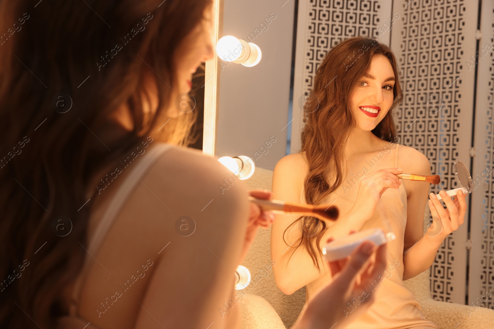 Photo of Beautiful young woman in elegant dress with powder and brush near mirror indoors