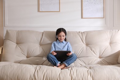 Photo of Little girl with headphones and tablet on sofa at home