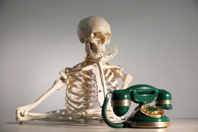 Waiting concept. Human skeleton at table with corded telephone against grey background