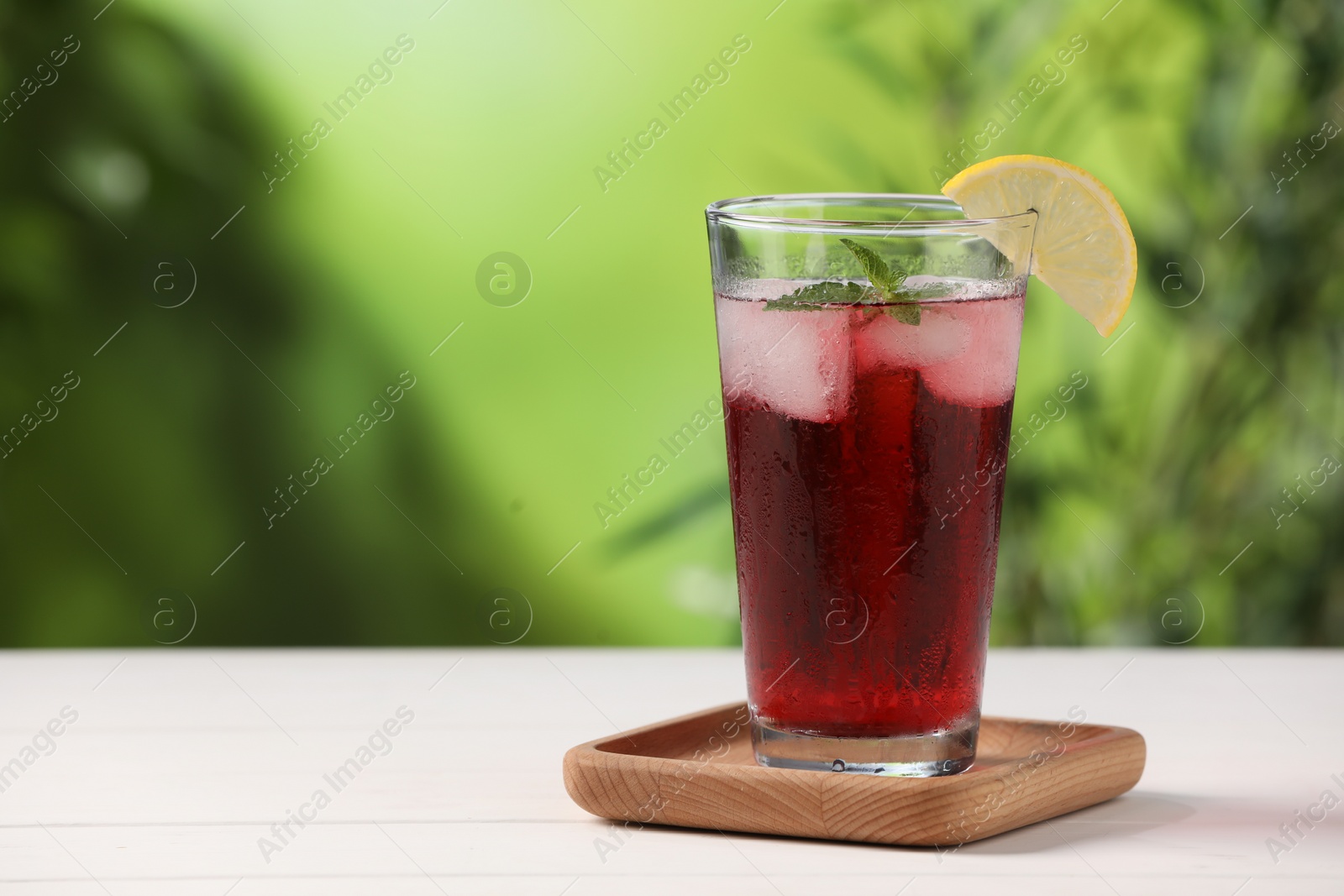 Photo of Refreshing hibiscus tea with ice cubes, mint and lemon in glass on white table against blurred green background. Space for text