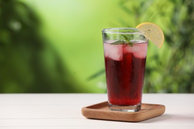 Photo of Refreshing hibiscus tea with ice cubes, mint and lemon in glass on white table against blurred green background. Space for text