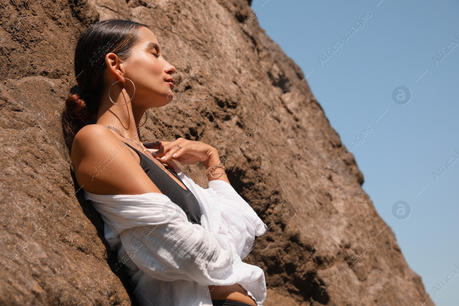 Photo of Beautiful young woman in stylish bikini near big stone outdoors on sunny day, space for text