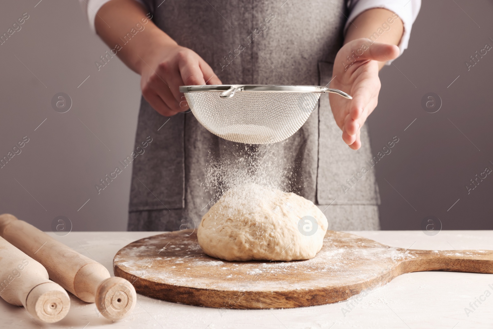 Photo of Man sprinkling flour over dough at table near grey wall, closeup