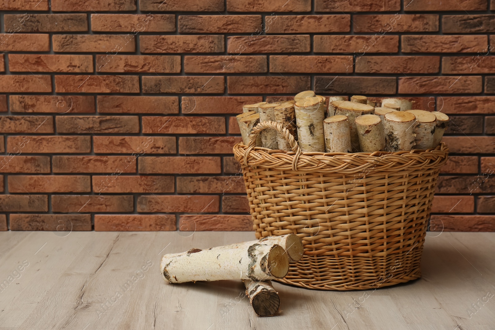 Photo of Wicker basket and firewood near brick wall indoors, space for text