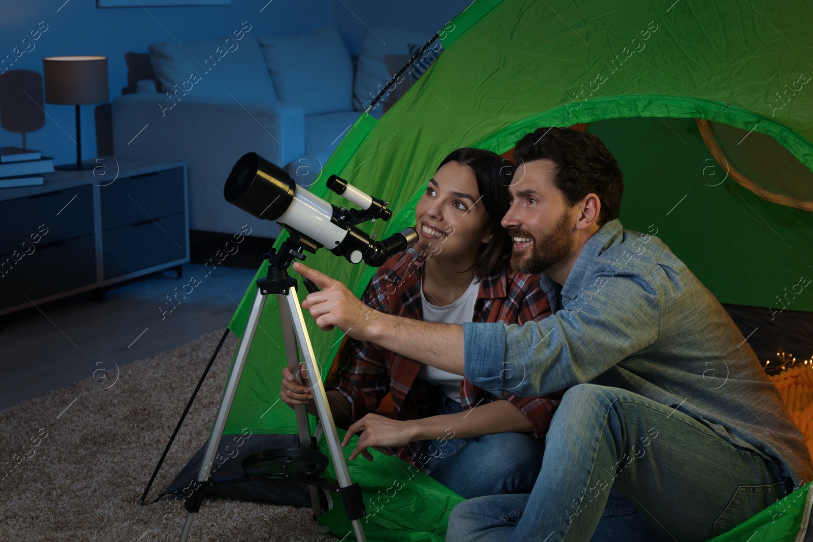 Photo of Happy couple using telescope to look at stars while sitting in camping tent indoors